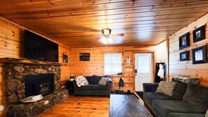 A cozy cabin interior with a stone fireplace, wooden walls, a ceiling fan, and a couch. - Six bedroom home with a hot tub, pool and an outdoor bar on a two acre lot. (Marquette)