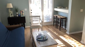 A living room with a blue sofa, a wooden coffee table with remote controls, a white chair, and a door with blinds. - Great Rates!  Screen Porch!  Pool Just Steps Away! (Seabrook Island)