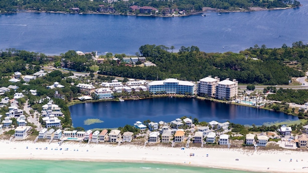 Ett flygbildsfoto av en kustort med en stor huvudbyggnad omgiven av mindre byggnader, en strand och en sjö.