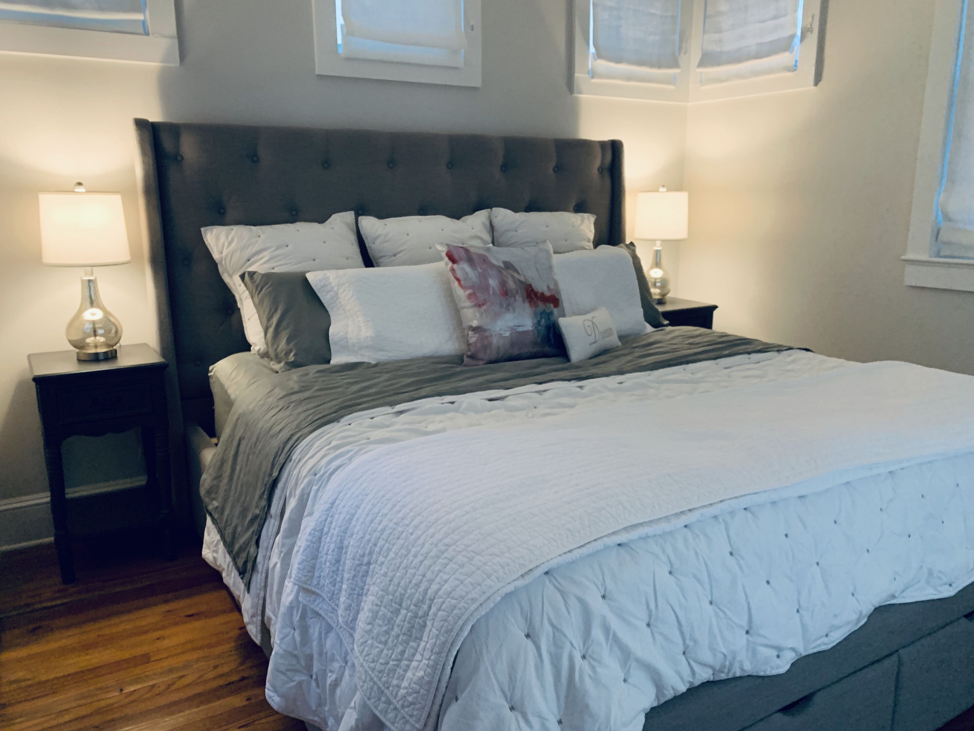 Una cama bien hecha con cabecera gris, ropa de cama blanca y varias almohadas. Hay dos mesitas de noche con lámparas, y el piso es de madera.