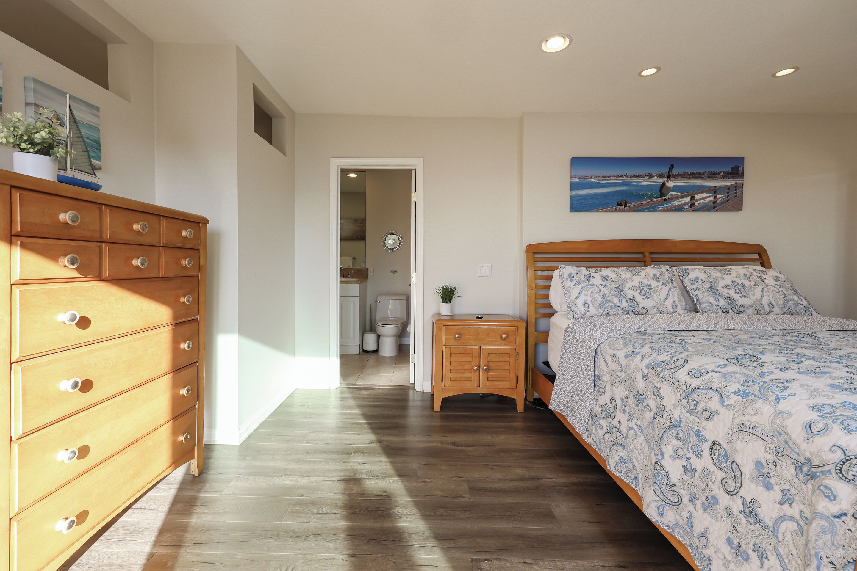 A bedroom with a wooden dresser, a bed with patterned bedding, a nightstand, and a painting on the wall.