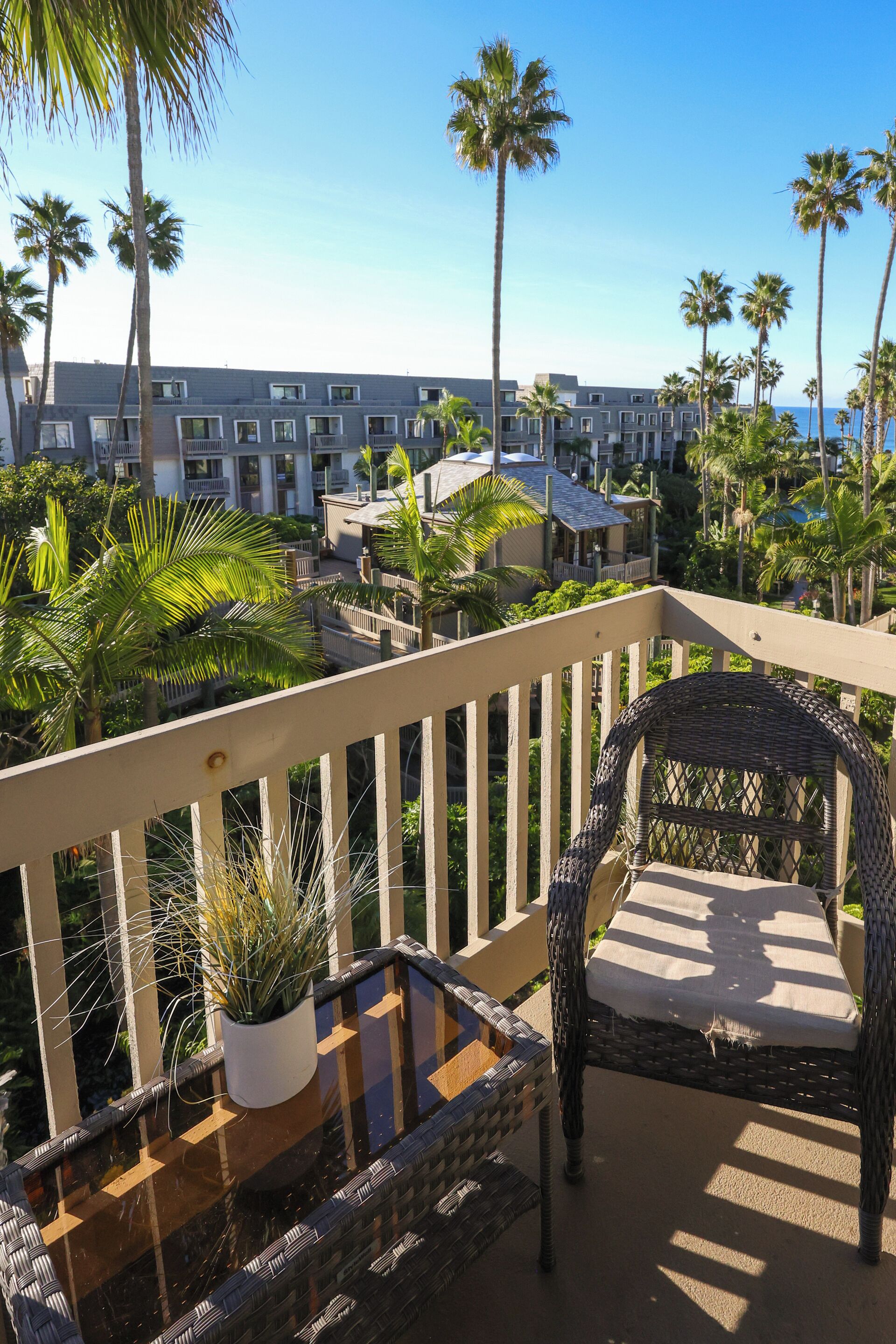 Een balkon met rieten meubilair, een potplant en uitzicht op een resort met palmbomen en gebouwen.