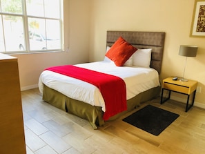 A neatly arranged bedroom with a bed, bedside table, lamp, and a window with a view of the outside.
