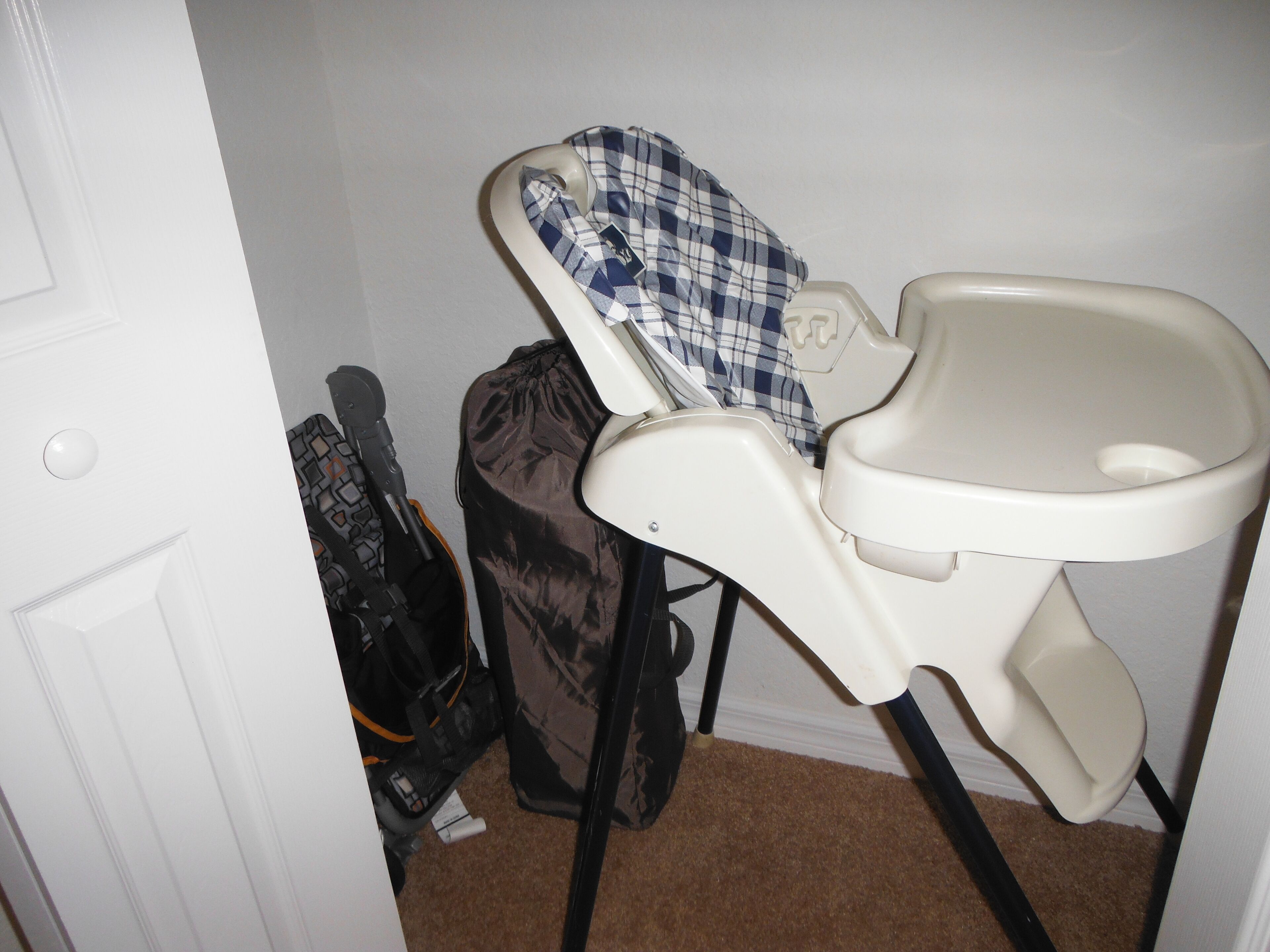 A baby high chair with a checkered cover, a black stroller, and a white door in a room with brown carpet.