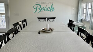 Uma área de refeições com uma mesa comprida, cadeiras pretas, toalha de mesa branca, uma vela e um prato decorativo com comida.