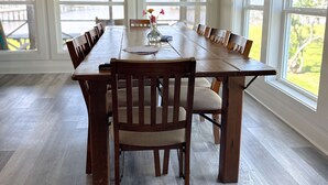 Una amplia mesa de comedor con múltiples sillas, ubicada en una habitación con ventanas grandes y suelo de madera.