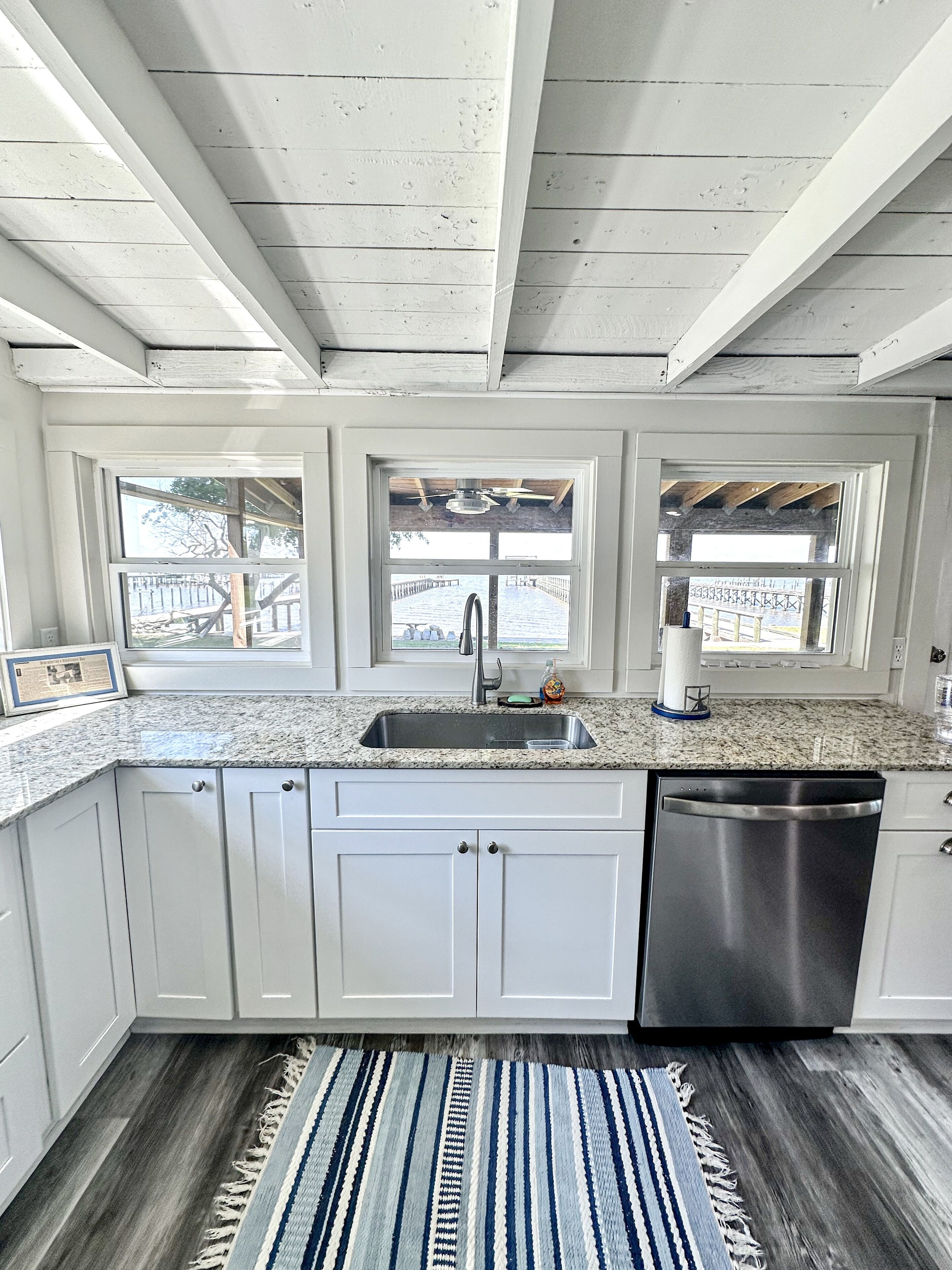 A kitchen with white cabinets, a stainless steel dishwasher, a granite countertop, and a view through large windows.