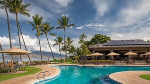 A pool area with lounge chairs and umbrellas, surrounded by palm trees and a view of the ocean.