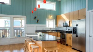 Une cuisine moderne dotée d’un îlot central, de tabourets en bois, d’appareils électroménagers en acier inoxydable et offrant une vue sur l’extérieur grâce à de grandes fenêtres.