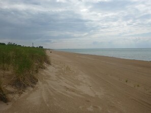 Ein weiter Sandstrand mit sanften Wellen, grasbewachsenen Dünen und einem fernen Leuchtturm.