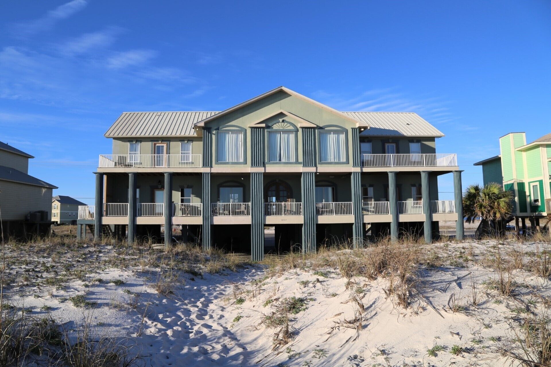 Una casa frente a la playa de dos pisos con un balcón en cada nivel, techo plano y una playa arenosa al frente.