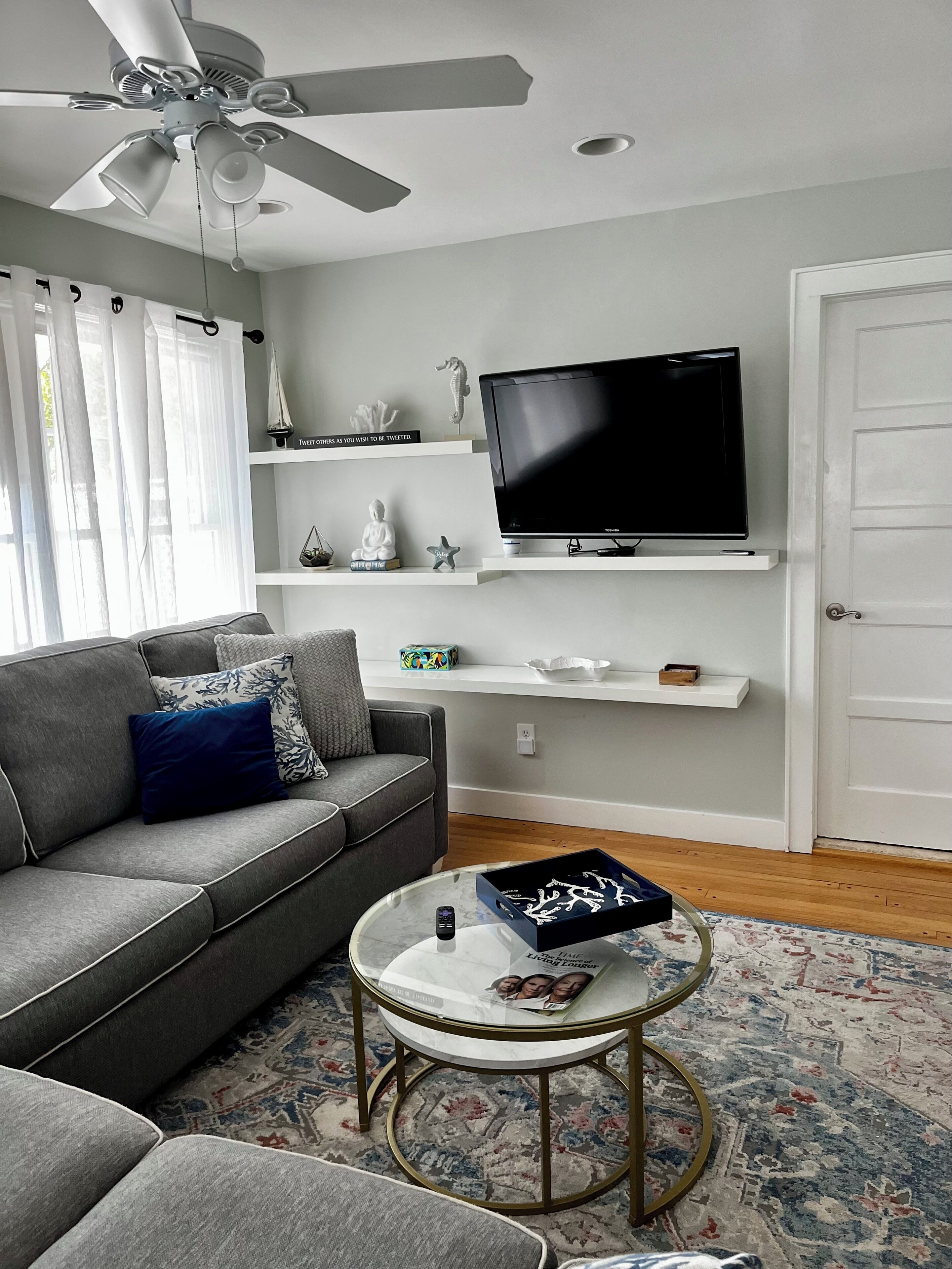 A living room with a grey sofa, a coffee table, a TV on a shelf, and a ceiling fan.