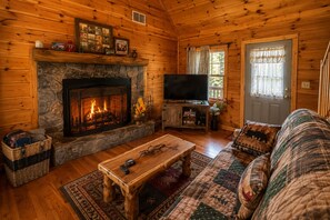 L’intérieur d’un chalet confortable, avec des murs en rondins de bois, une cheminée en pierre, un téléviseur, une table basse en bois et un canapé à motifs.
