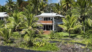 A two-story house surrounded by lush tropical vegetation, including palm trees and various shrubs. - Luxuriously Appointed Bali Temple Has 160 Degree Ocean Views! (Pahoa)
