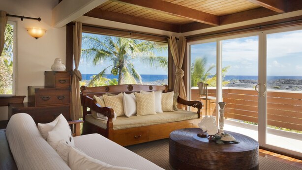 A bedroom with a bed, a wooden bench, a nightstand, and a window with a view of the ocean and palm trees. - Luxuriously Appointed Bali Temple Has 160 Degree Ocean Views! (Pahoa)