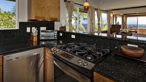A modern kitchen with a dishwasher, stove, and oven, wooden cabinets, and a view of the ocean. - Luxuriously Appointed Bali Temple Has 160 Degree Ocean Views! (Pahoa)
