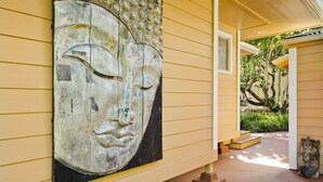 A building exterior with a large Buddha statue on the wall, a covered porch, and a small garden area. - Luxuriously Appointed Bali Temple Has 160 Degree Ocean Views! (Pahoa)