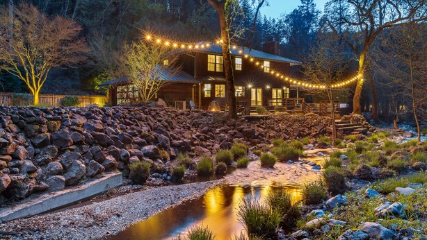 Una cabaña rústica con un muro de contención de piedra, un pequeño arroyo y luces colgantes.