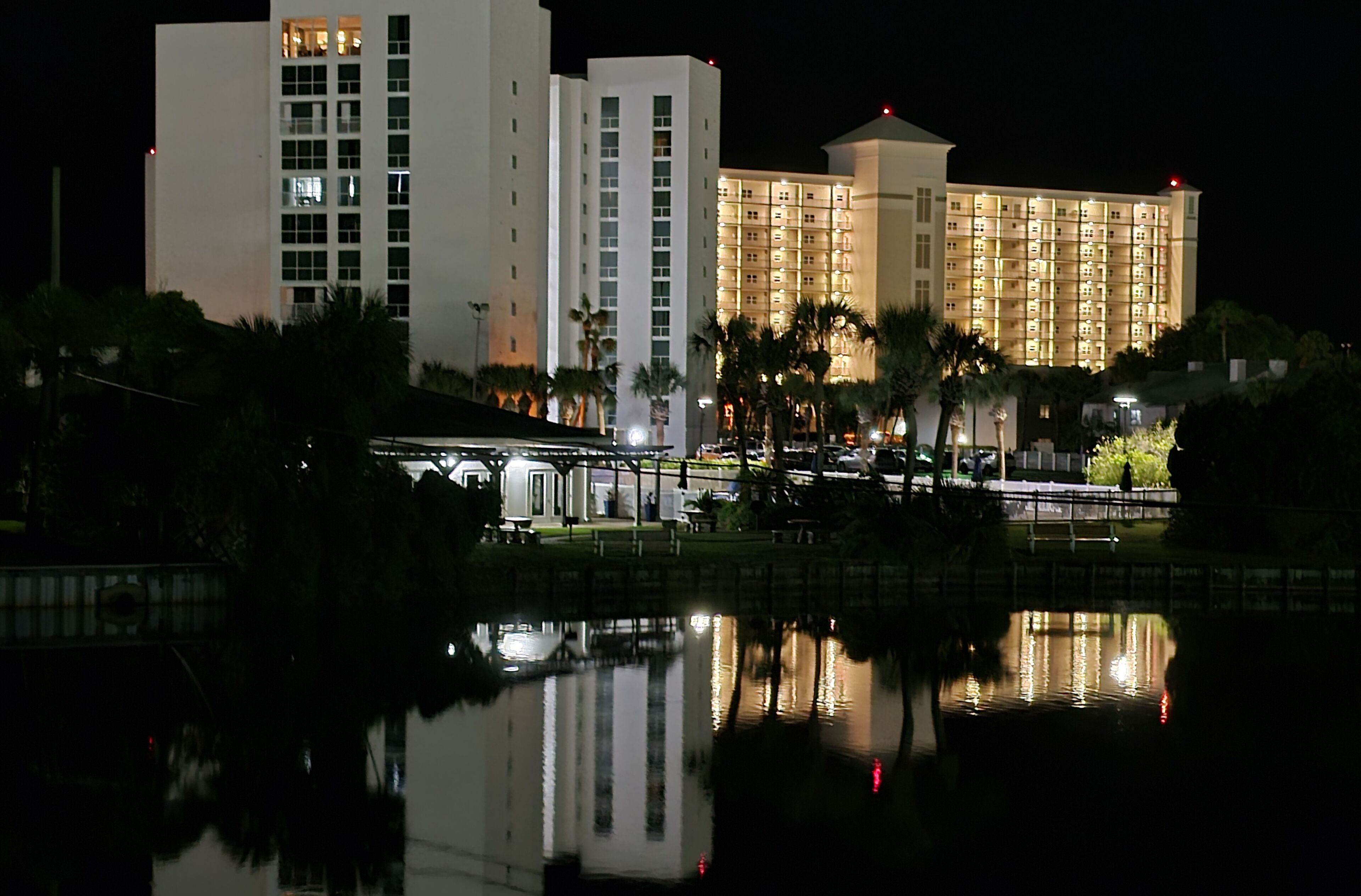 A hotel with multiple high-rise buildings, a reflective water body in the foreground, and palm trees.