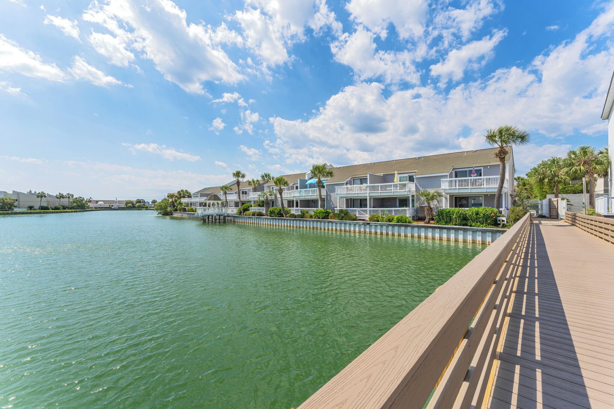 A waterfront property with a wooden deck, a pool, and a building with balconies and palm trees.