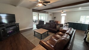 A living room with a TV, leather sofa, coffee table, and a ceiling fan. - Louisiana Tickfaw River House Near New Orleans, Baton Rouge! Best Views! HotTub! (Springfield)