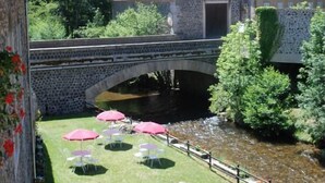 Uma ponte de pedra sobre um rio, rodeada por vegetação exuberante e um edifício de pedra.