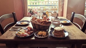 Mesa de café da manhã com pão, doces e café, em frente a uma janela com vista para prédios e árvores.