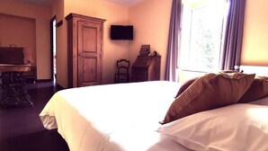 Quarto de hotel com uma cama grande, um guarda-roupa de madeira, uma escrivaninha com cadeira e uma televisão fixada na parede.