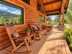 A log cabin with a porch featuring rocking chairs, a table, and a view of a forested landscape. - Luxury lodging minutes from Glacier National Park. (East Glacier Park)