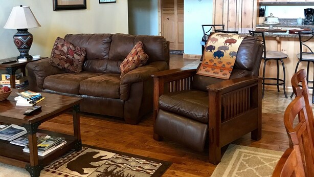 A cozy living room with a brown leather sofa, a patterned armchair, and a coffee table with books and a lamp. A rug with animal prints covers the wooden floor.