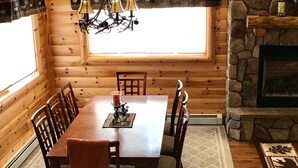 A wooden interior with a dining table, chairs, a chandelier, a fireplace, and a window.