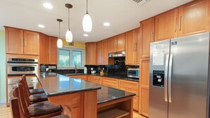 A modern kitchen with wooden cabinets, a stainless steel refrigerator, a built-in oven, and a large island with bar stools. - Sunbeam-Soundview Private Home. 2 Min Walk to the Beach (Navarre)