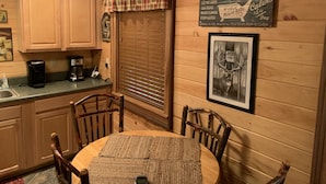 A cozy lodge kitchen with wooden cabinets, a plaid window valance, a framed deer picture, and a sign advertising amenities like electric lights and hot mineral baths.
