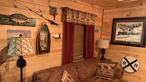 A cozy cabin interior with a brown couch, a lamp, a framed picture, and a 'Big Catch' sign.