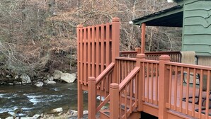 A wooden deck with a staircase leading to a covered area, overlooking a river with rocks and trees in the background.