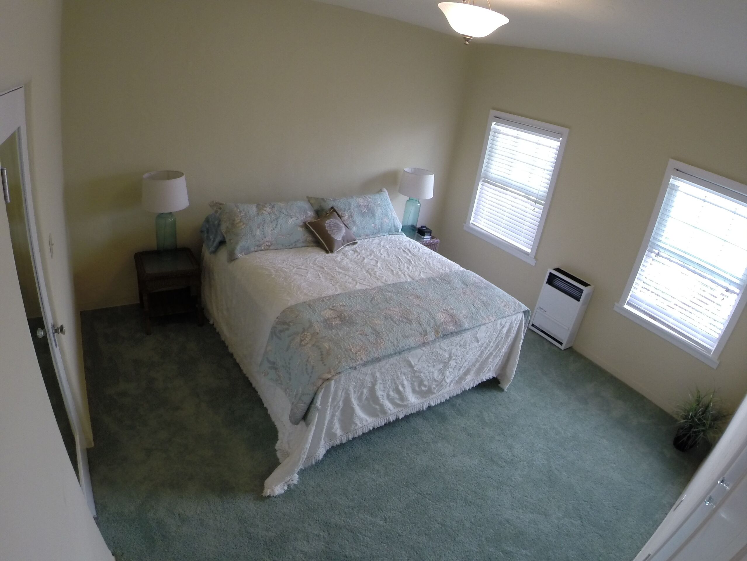 A bedroom with a large bed, two bedside lamps, a heater, and two windows with blinds.