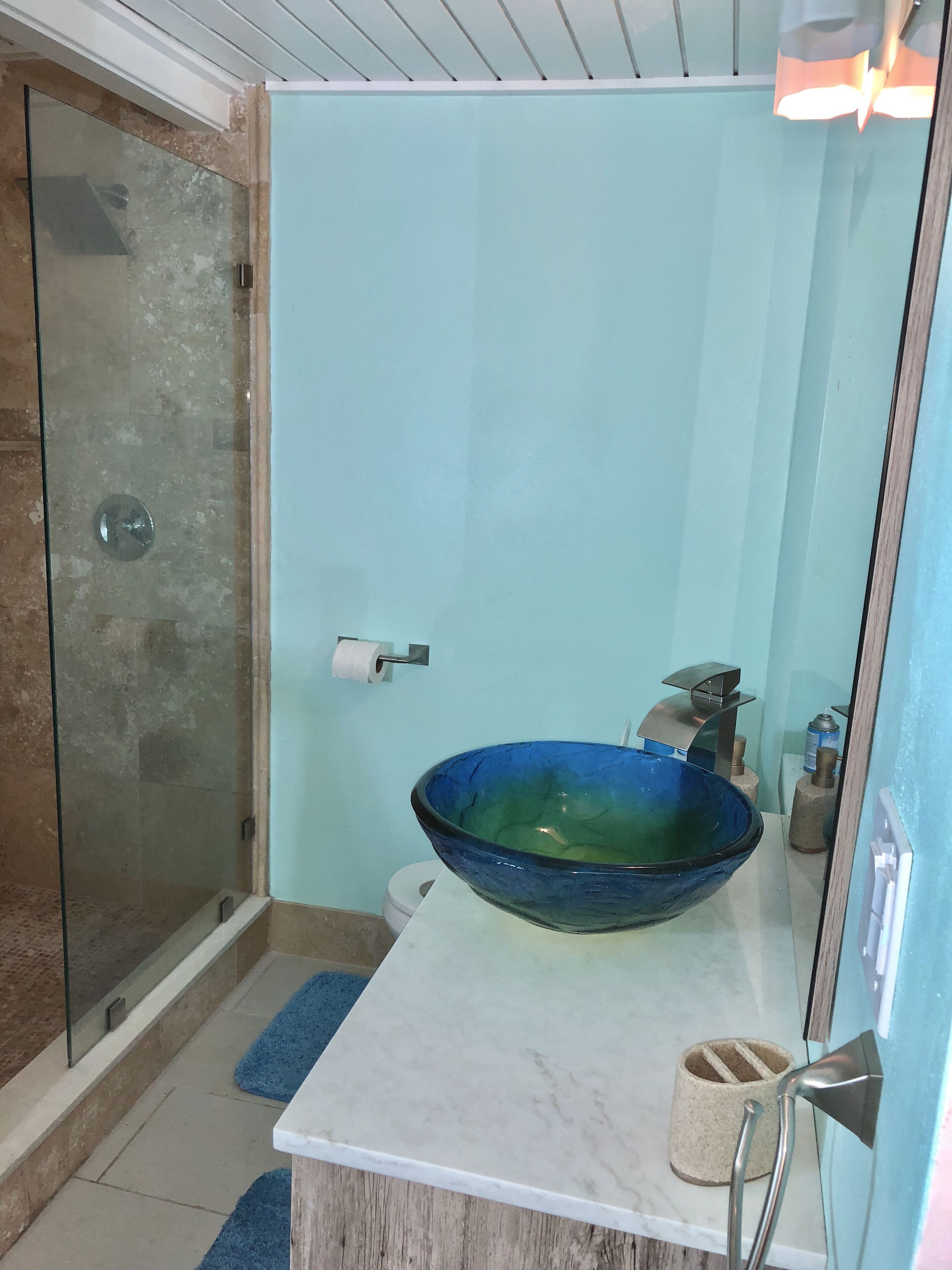 Un baño con cabina de ducha de vidrio, lavabo de vidrio azul, encimera de mármol blanco y un perchero para toallas.