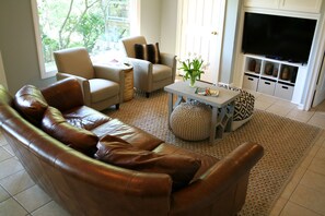 Uma sala de estar com um sofá de couro marrom, poltronas bege, uma mesa de centro e uma TV sobre um suporte.