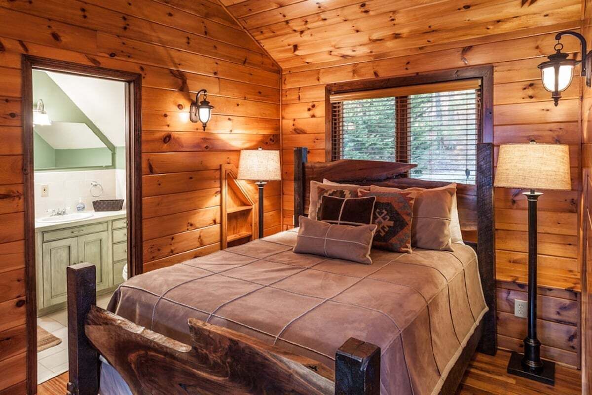 A wooden cabin bedroom with a bed, two bedside lamps, and a window with blinds.