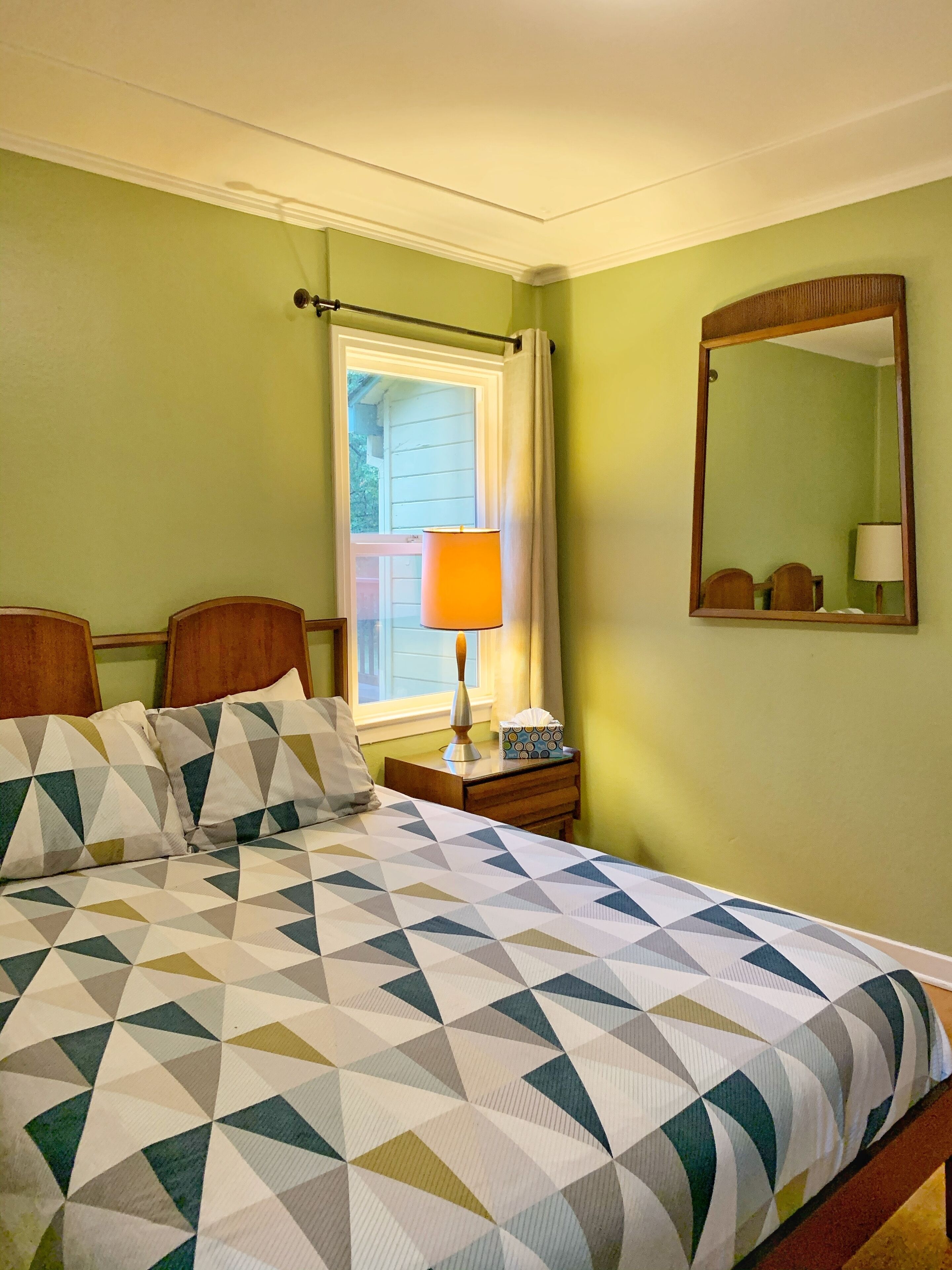 Une chambre d’hôtel avec un lit, une table de chevet, une lampe, une fenêtre avec des rideaux et un miroir au mur.