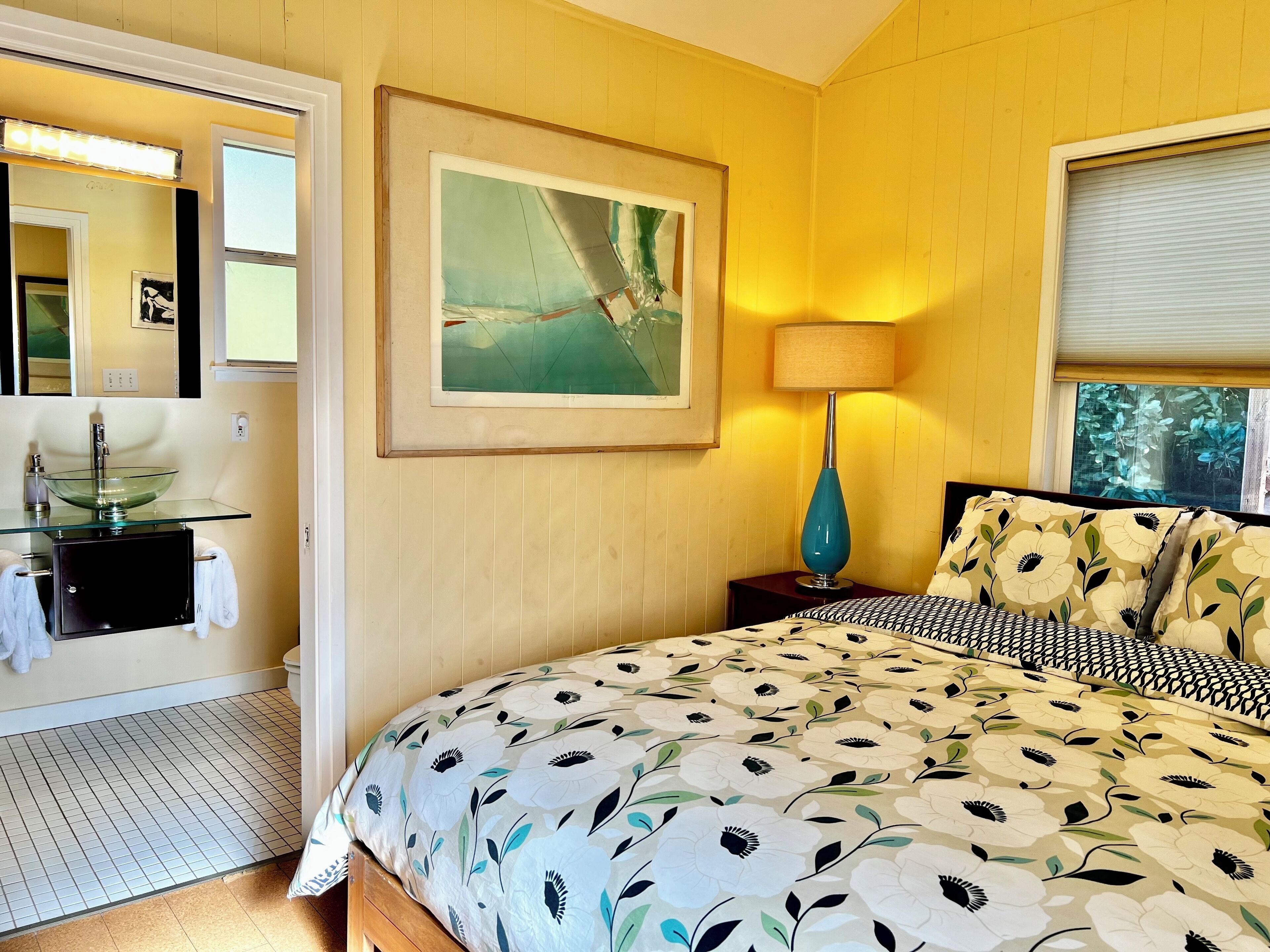 A bedroom with a bed, a bedside table with a lamp, a framed painting, a bathroom with a sink and mirror, and a window with blinds.