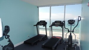 Une salle de remise en forme équipée de tapis de course, d’un vélo elliptique et d’un vélo d’appartement, avec vue sur la plage grâce à de grandes fenêtres.