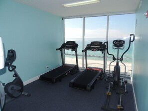 Une salle de remise en forme équipée de tapis de course, d’un vélo elliptique et d’un vélo d’appartement, avec vue sur la plage grâce à de grandes fenêtres.