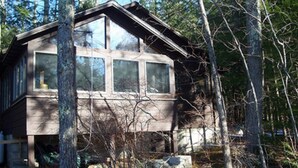 A cabin with a simple, rectangular structure, large windows, and a sloped roof, surrounded by trees. - Lake Front Cabin on Squam Lake/On Golden Pond (Center Harbor)