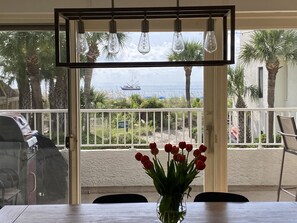 A dining area with a table, chairs, a vase of red flowers, and a view of the ocean through a glass door.