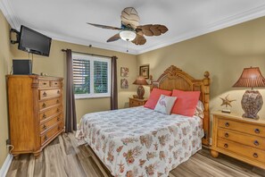A bedroom with a bed, wooden dresser, nightstand, lamp, ceiling fan, TV, and window with blinds.