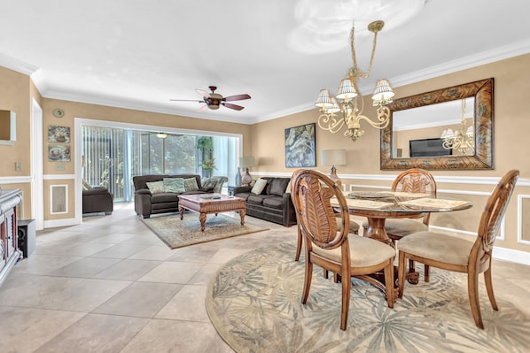 A spacious living room with a large window, a ceiling fan, a chandelier, and a dining area with a round table and chairs.
