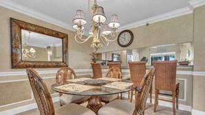 Uma área de jantar com uma mesa redonda de vidro, cadeiras de vime, um candelabro e um espelho grande na parede.