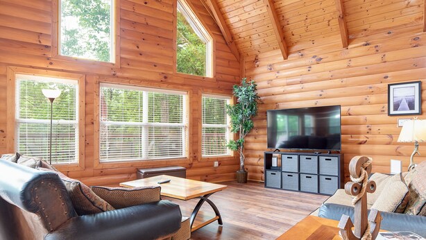 Ein Holzblockhaus-Interieur mit Holzbalkendecke, großen Fenstern, einem Flachbildfernseher und einem Essbereich.