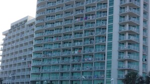 A high-rise hotel with multiple balconies, a sandy beach in the foreground, and a fenced area with palm trees. - Myrtle Beach Oceanfront Condo. Sept. open Dates 79 a night. (Myrtle Beach)